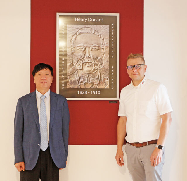 Vizerektor des Gansu Medical Colleges Yumin Zhao und Markus Ruckdeschel, Geschäftsführer des BRK-Kreisverbandes Bayreuth,vor einem Bild von Henry Dunant - Gründer der Rot-Kreuz-Bewegung.